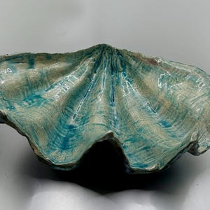 May include: A ceramic clam shell dish with a teal and white glaze. The shell is open and has a smooth, rounded interior.