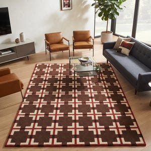 Puede incluir: Una alfombra rectangular con un patrón geométrico repetitivo de cuadrados de color marrón oscuro acentuados con motivos en forma de cruz en blanco y rojo. La alfombra está extendida sobre un suelo de madera de tono claro.