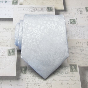 以下が含まれることがあります： 白い花柄の薄いブルーのネクタイ。ネクタイは絹のような素材で、上品な光沢があります。