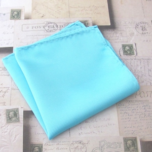 May include: A folded, light blue pocket square with a ribbed texture. The square is made of a soft fabric and is neatly folded, showcasing its clean lines and vibrant color. The background features vintage postcards.