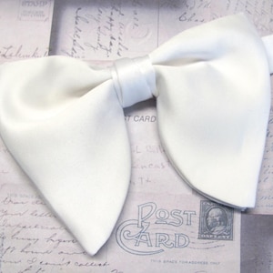 May include: A pristine white bow tie, likely satin or silk, is centered in the frame. The bow tie is neatly tied, with a classic butterfly shape. The background features vintage postcard imagery.