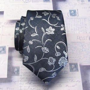 Peut inclure: Une cravate noire ornée d'un motif floral argenté répétitif avec des tiges et des feuilles entrelacées. Le tissu présente une finition légèrement texturée, et la cravate est pliée pour montrer clairement l'extrémité pointue et le motif.