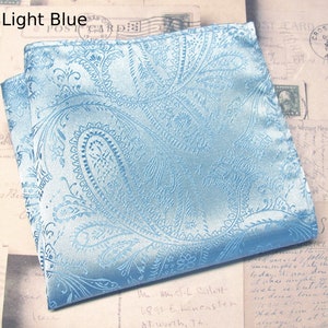 May include: A light blue paisley pocket square, folded to display its intricate design. The fabric has a sheen, and the pattern is a repeating floral motif. The text "Light Blue" is visible. The pocket square is a classic accessory.