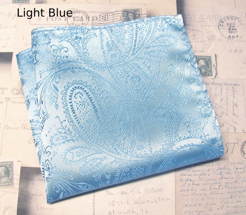 May include: A light blue paisley pocket square, folded to display its intricate design. The fabric has a sheen, and the pattern is a repeating floral motif. The text "Light Blue" is visible. The pocket square is a classic accessory.