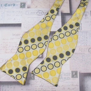 May include: A bow tie with a white background and a pattern of yellow and black circles. The bow tie is made of a silky material.