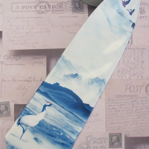 May include: A light blue silk tie with a watercolor design of a mountain range, a lake, and a crane. The tie is on a background of vintage postcards.