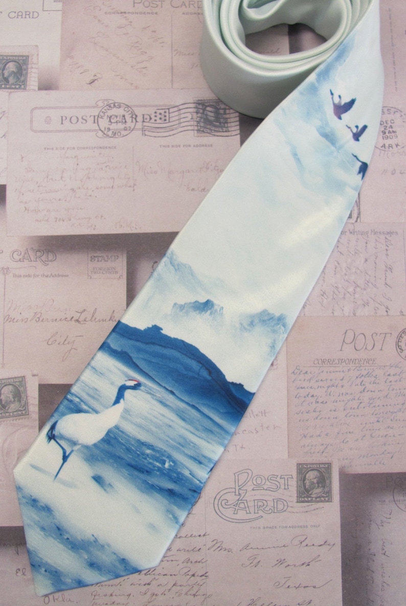 May include: A light blue silk tie with a watercolor design of a mountain range, a lake, and a crane. The tie is on a background of vintage postcards.