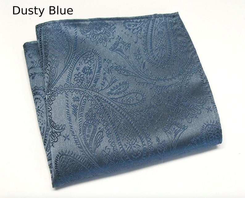 May include: A folded dusty blue paisley pocket square. The fabric has a subtle sheen and a detailed, repeating pattern. The text "Dusty Blue" is in the upper left corner. This accessory is suitable for formal wear.