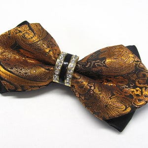 May include: A black bow tie with a gold and black patterned design. The bow tie has a decorative ring with clear rhinestones.