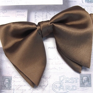 Könnte beinhalten: Eine braune Schleife aus Satin. Die Schleife ist einfarbig und hat eine klassische Form. Der Hintergrund ist ein Vintage-Postkarten-Design.