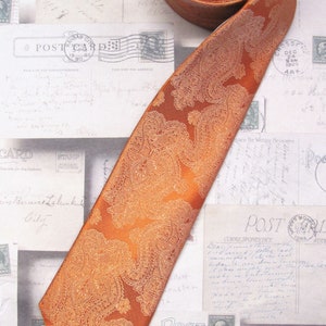 Pu&ograve; includere: Una cravatta di colore arancio bruciato con motivo paisley su sfondo bianco. La cravatta &egrave; piegata e appoggiata su una superficie con cartoline d'epoca.