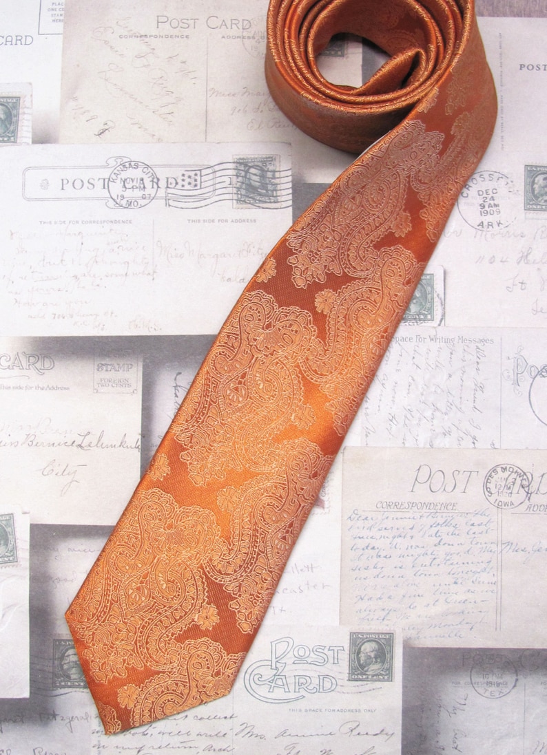 Pu&ograve; includere: Una cravatta di colore arancio bruciato con motivo paisley su sfondo bianco. La cravatta &egrave; piegata e appoggiata su una superficie con cartoline d'epoca.
