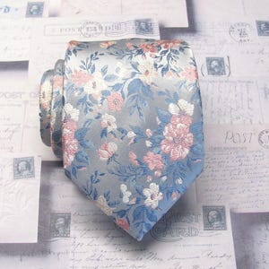 Peut inclure: Une cravate présentant une base bleu clair avec un motif floral tissé dans des tons de rose, blanc et bleu plus foncé. Le tissu a un éclat subtil et le motif se compose de diverses fleurs épanouies et de tiges feuillues sur la surface.