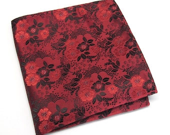 May include: A folded red pocket square with a floral pattern. The design features red flowers and black leaves and stems against a red background. The pocket square is made of a shiny, silky material.