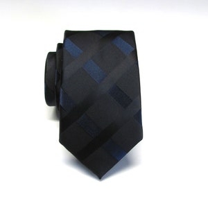 May include: A black tie featuring a repeating pattern of dark blue and black rectangles and stripes. The tie is rolled up, angled towards the viewer, displaying the design. The background is plain white.