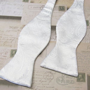 May include: A white bow tie with a paisley pattern. The bow tie is made of a satin fabric and has a smooth texture.
