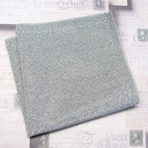 May include: Two folded, square, silver-grey fabric napkins. The napkins have a textured, woven appearance with a subtle shimmer. The background features vintage postcards with handwritten text and postage stamps.