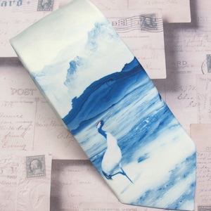 May include: A white silk tie with a blue and white watercolor design of a crane standing in a body of water with mountains in the background.