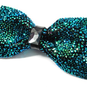 May include: A teal and black bow tie covered in sparkling crystals. The bow tie has a black leather-like band in the center with a large, clear, rectangular crystal. The bow tie is on a white background.
