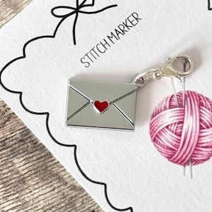 Op de afbeelding: Een zilveren steekmarkeerder in de vorm van een envelop met een rood hart, bevestigd aan een roze garenbalbedel. De steekmarkeerder staat op een witte kaart met de woorden "STITCH MARKER" en een zwarte strik.