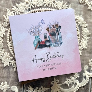 Könnte beinhalten: Rosa Geburtstagskarte mit einer Aquarellillustration von Make-up-Pinseln, einer Vase mit Blumen und einem Schmetterling. Der Text lautet "Happy Birthday TO A VERY SPECIAL DAUGHTER".