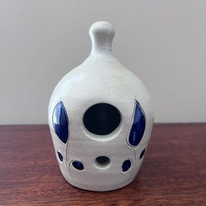 May include: A bell-shaped ceramic object with a matte off-white finish, featuring a narrow neck and a rounded body. The surface is decorated with carved, glossy dark blue leaf-shaped accents and includes a large circular opening near the top.