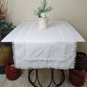May include: White table runner with a frayed edge, placed on a white table with a black metal base. A white pitcher with a bouquet of white flowers sits on the table runner.