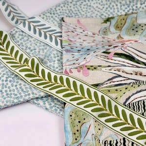 May include: Close-up of various fabric samples and decorative ribbons. The ribbons feature a white background with green leaf designs. Other fabrics include a blue dotted pattern and a floral print with green, blue, and pink hues.