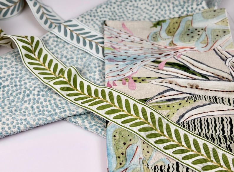May include: Close-up of various fabric samples and decorative ribbons. The ribbons feature a white background with green leaf designs. Other fabrics include a blue dotted pattern and a floral print with green, blue, and pink hues.