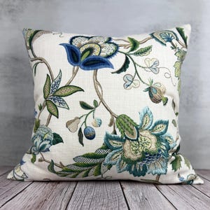May include: Square decorative pillow with a cream-colored background and a floral pattern in shades of blue, green, and brown. The pillow features embroidered flowers and leaves, creating a botanical design. The pillow is placed on a wooden surface.