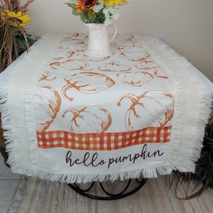 May include: A white burlap table runner with a frayed edge features a pattern of orange pumpkins and a red and white checkered stripe. The text "hello pumpkin" is printed in brown on the runner.