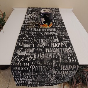 May include: Black and white Halloween themed table runner with a white background and black text. The text includes words like "Halloween", "Hauntings", "Potions", "Cobwebs", "Spiders", "Jack-O-Lantern", "31st", "Oct", and "Happy".