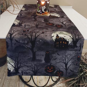 May include: Black and gray Halloween table runner with a spooky haunted house, black cats, pumpkins, bats, and trees. The runner is on a white table with a black metal base.