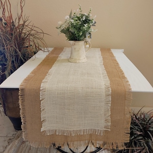 May include: A white and brown burlap table runner with frayed edges. The runner is on a white table with a white pitcher filled with green and white flowers.