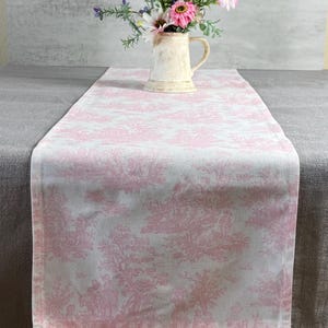 May include: A pink and white toile table runner with a floral arrangement in a cream-colored pitcher. The runner features a repeating pattern of pastoral scenes. The flowers include pink, white, and purple blooms.