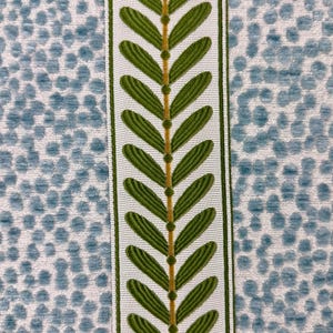 May include: Close-up of a decorative fabric with a repeating pattern. The design features a central vertical stripe with green leaf-like shapes on a white background, bordered by green trim. The background is a light blue dotted pattern.