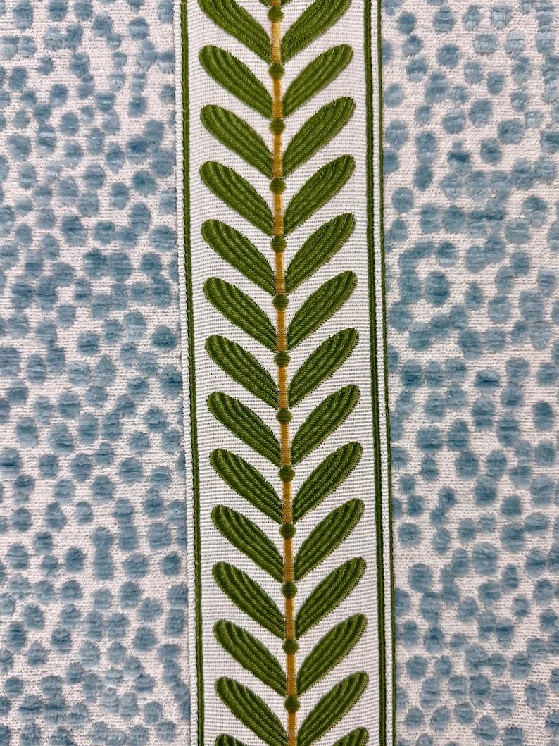 May include: Close-up of a decorative fabric with a repeating pattern. The design features a central vertical stripe with green leaf-like shapes on a white background, bordered by green trim. The background is a light blue dotted pattern.