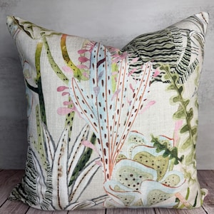 K&ouml;nnte beinhalten: Quadratisches Zierkissen mit botanischem Druck. Das Kissen hat einen cremefarbenen Hintergrund und ein farbenfrohes Design aus Pflanzen in Gr&uuml;n-, Rosa- und Blaut&ouml;nen. Das Kissen ist aus einem strukturierten Stoff gefertigt.