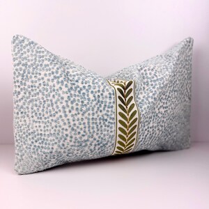 May include: A decorative pillow with a light blue and white speckled pattern. A vertical band of cream-colored fabric with embroidered green leaves accents the center. The pillow is rectangular and appears to be made of a textured fabric.