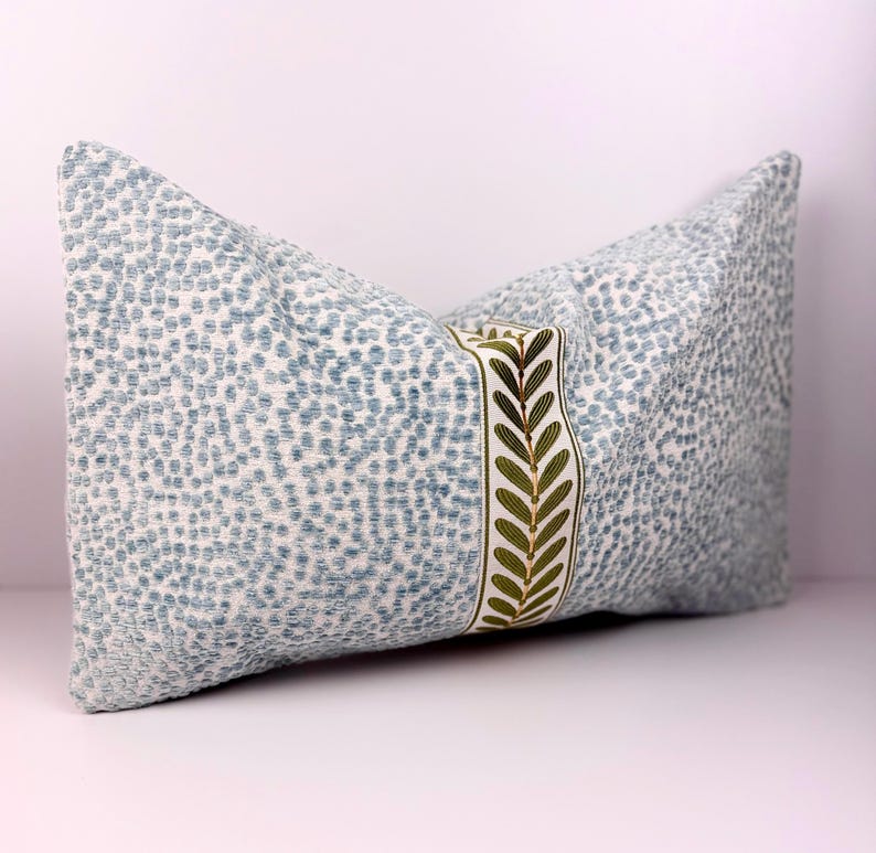 May include: A decorative pillow with a light blue and white speckled pattern. A vertical band of cream-colored fabric with embroidered green leaves accents the center. The pillow is rectangular and appears to be made of a textured fabric.