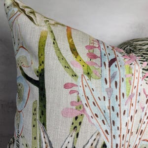 K&ouml;nnte beinhalten: Nahaufnahme eines dekorativen Kissens mit botanischem Druck. Das Kissen hat einen hellbeigen Hintergrund mit einem farbenfrohen Design aus gr&uuml;nen, rosa und blauen Pflanzenillustrationen. Die Ecke des Kissens ist sichtbar und zeigt die Textur des Stoffes.