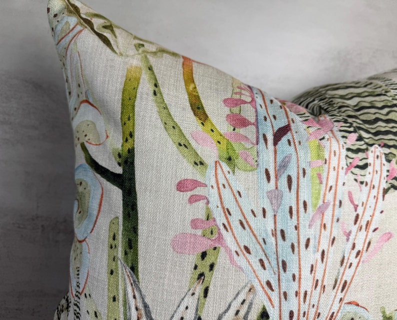 K&ouml;nnte beinhalten: Nahaufnahme eines dekorativen Kissens mit botanischem Druck. Das Kissen hat einen hellbeigen Hintergrund mit einem farbenfrohen Design aus gr&uuml;nen, rosa und blauen Pflanzenillustrationen. Die Ecke des Kissens ist sichtbar und zeigt die Textur des Stoffes.