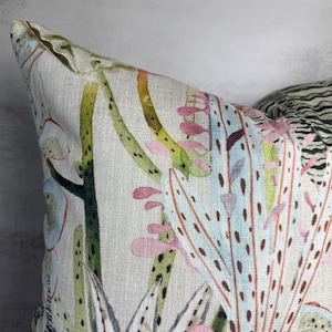 K&ouml;nnte beinhalten: Nahaufnahme eines dekorativen Kissens mit botanischem Druck. Das Kissen hat einen hellbeigen Hintergrund mit einem Aquarelldesign von Kakteen und anderen W&uuml;stenpflanzen in Gr&uuml;n-, Rosa- und Blaut&ouml;nen. Das Kissen hat ein strukturiertes Aussehen.