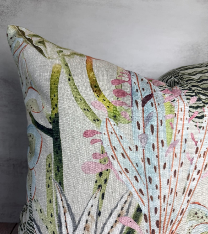 K&ouml;nnte beinhalten: Nahaufnahme eines dekorativen Kissens mit botanischem Druck. Das Kissen hat einen hellbeigen Hintergrund mit einem Aquarelldesign von Kakteen und anderen W&uuml;stenpflanzen in Gr&uuml;n-, Rosa- und Blaut&ouml;nen. Das Kissen hat ein strukturiertes Aussehen.