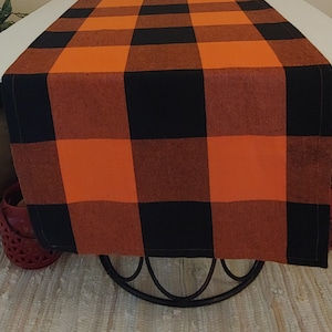 May include: Orange and black checkered table runner. The runner is made of a woven fabric and has a rustic look.