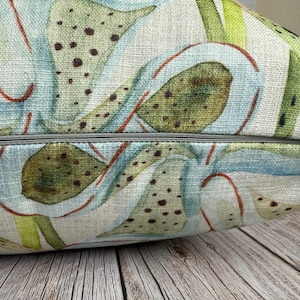 K&ouml;nnte beinhalten: Ein dekoratives Kissen mit botanischem Design. Das Kissen zeigt einen Aquarelldruck von gr&uuml;nen, blauen und braunen pflanzenartigen Formen auf hellem beigen Hintergrund. Ein grauer Rei&szlig;verschluss ist am unteren Rand sichtbar.