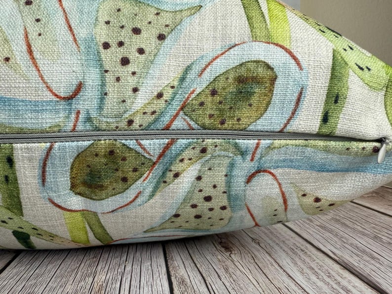 K&ouml;nnte beinhalten: Ein dekoratives Kissen mit botanischem Design. Das Kissen zeigt einen Aquarelldruck von gr&uuml;nen, blauen und braunen pflanzenartigen Formen auf hellem beigen Hintergrund. Ein grauer Rei&szlig;verschluss ist am unteren Rand sichtbar.