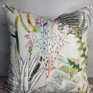 K&ouml;nnte beinhalten: Quadratisches Zierkissen mit botanischem Druck. Das Kissen hat einen hellen Hintergrund mit einem farbenfrohen Design verschiedener Pflanzen in Gr&uuml;n-, Rosa- und Blaut&ouml;nen. Das Kissen liegt auf einer Holzoberfl&auml;che.