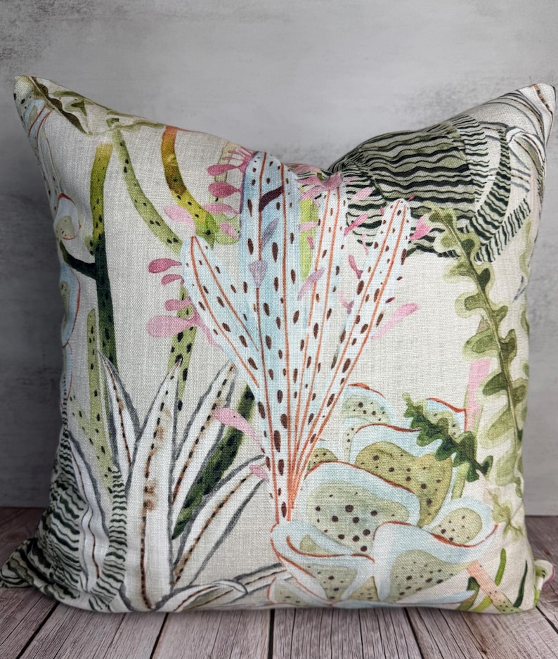 K&ouml;nnte beinhalten: Quadratisches Zierkissen mit botanischem Druck. Das Kissen hat einen hellen Hintergrund mit einem farbenfrohen Design verschiedener Pflanzen in Gr&uuml;n-, Rosa- und Blaut&ouml;nen. Das Kissen liegt auf einer Holzoberfl&auml;che.