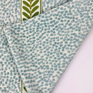May include: Close-up of a textured fabric with a repeating pattern of small, light blue dots on a white background. A folded section reveals the fabric's texture. A vertical stripe with green leaf designs on a white background adds a decorative element.